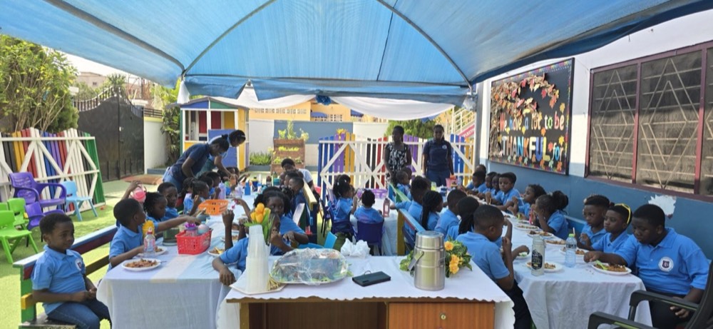 Pupils in blue uniform seated along a long table for a shared meal outdoors